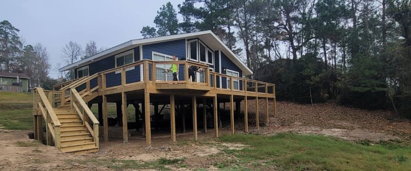 Wide deck with stairs built onto a raised blue-gray house on piers