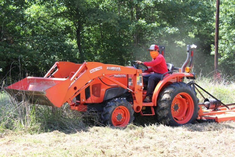 Tractor with a front loader and rear rotary cutter mowing overgrown grass